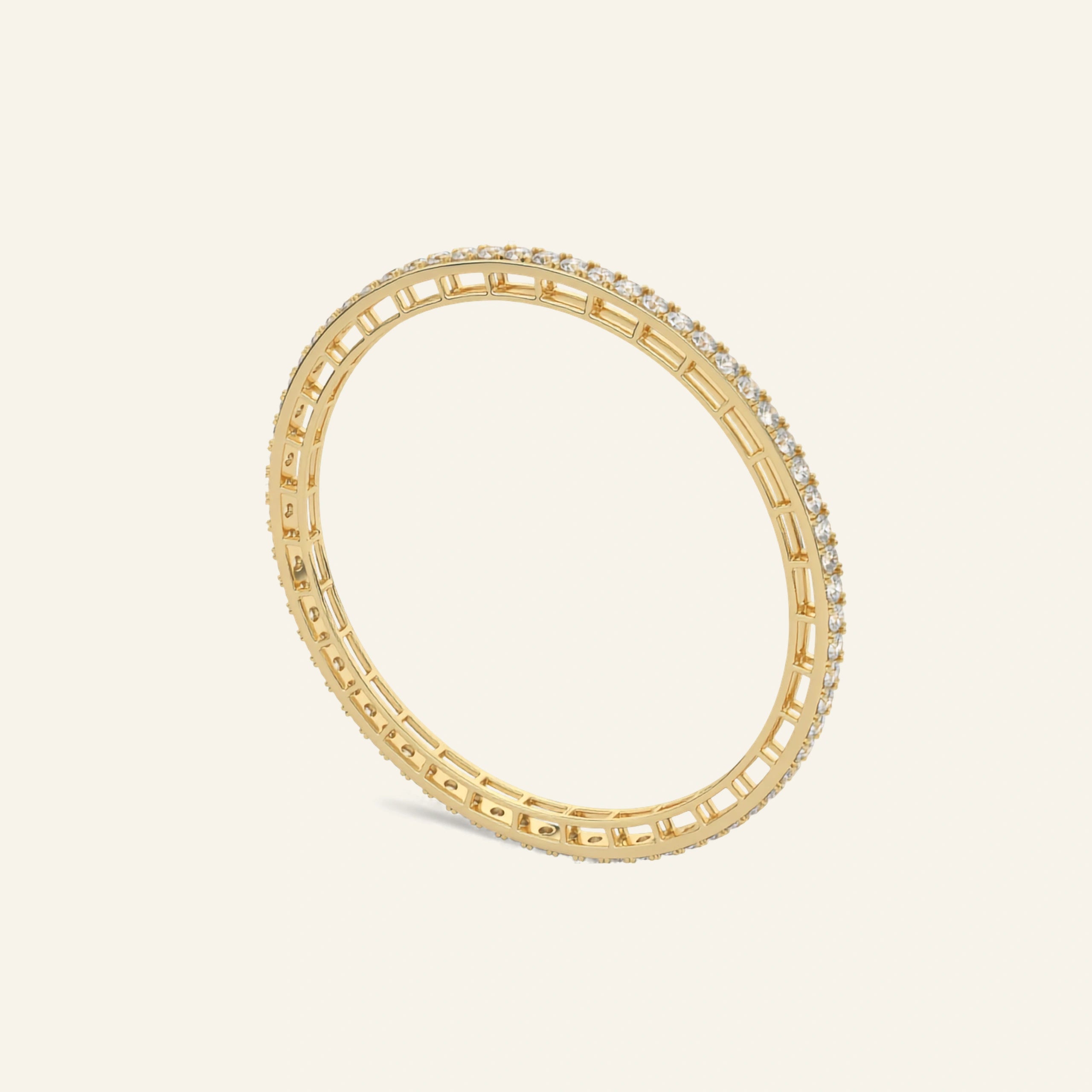 A gold bangle bracelet featuring a continuous row of small clear stones along one side, displayed on a light cream background.